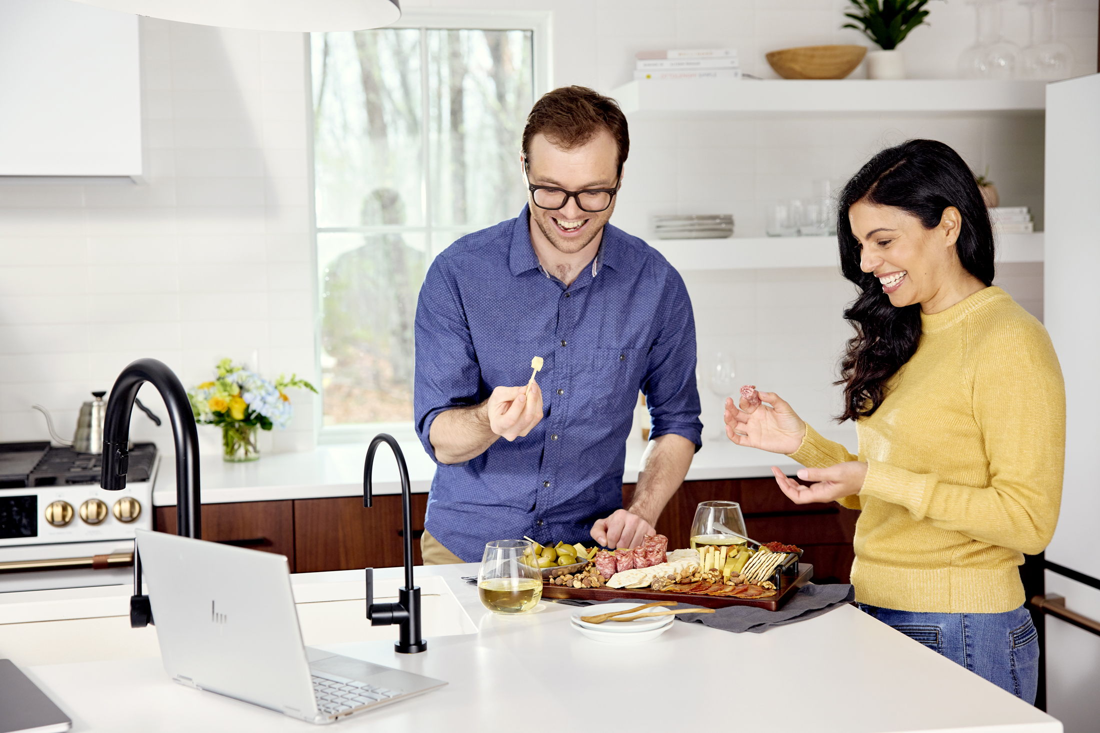 Two epople in a kitchen making a charcuterie board and having wine while following an Alice's Table Virtual Workshop on a laptop