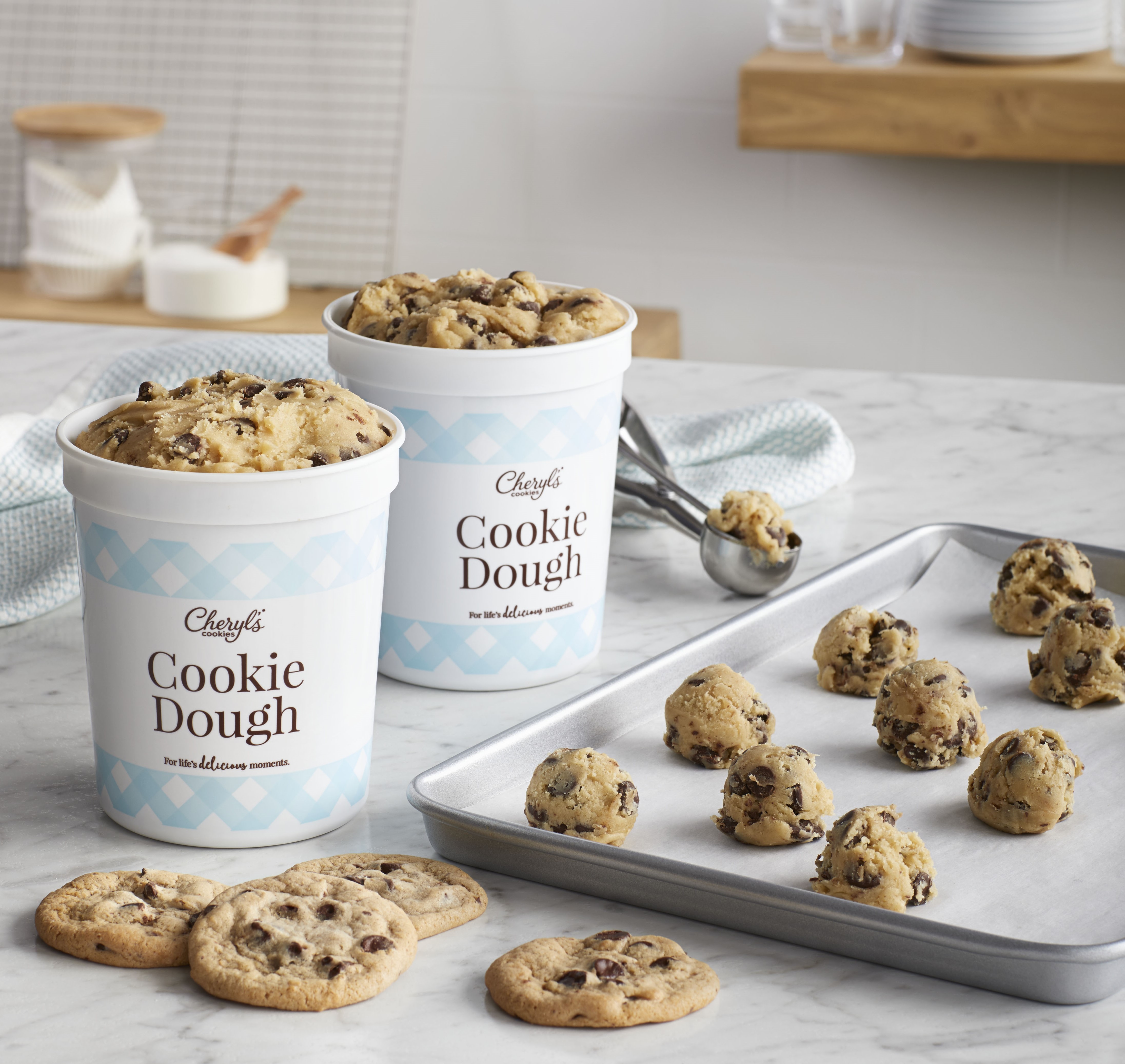 Two tubs of Cheryl's Cookies Chocolate Chip Bake-at-Home Cookie Dough next to a baking tray of cookie dough balls in rows and some already baked cookies on the side