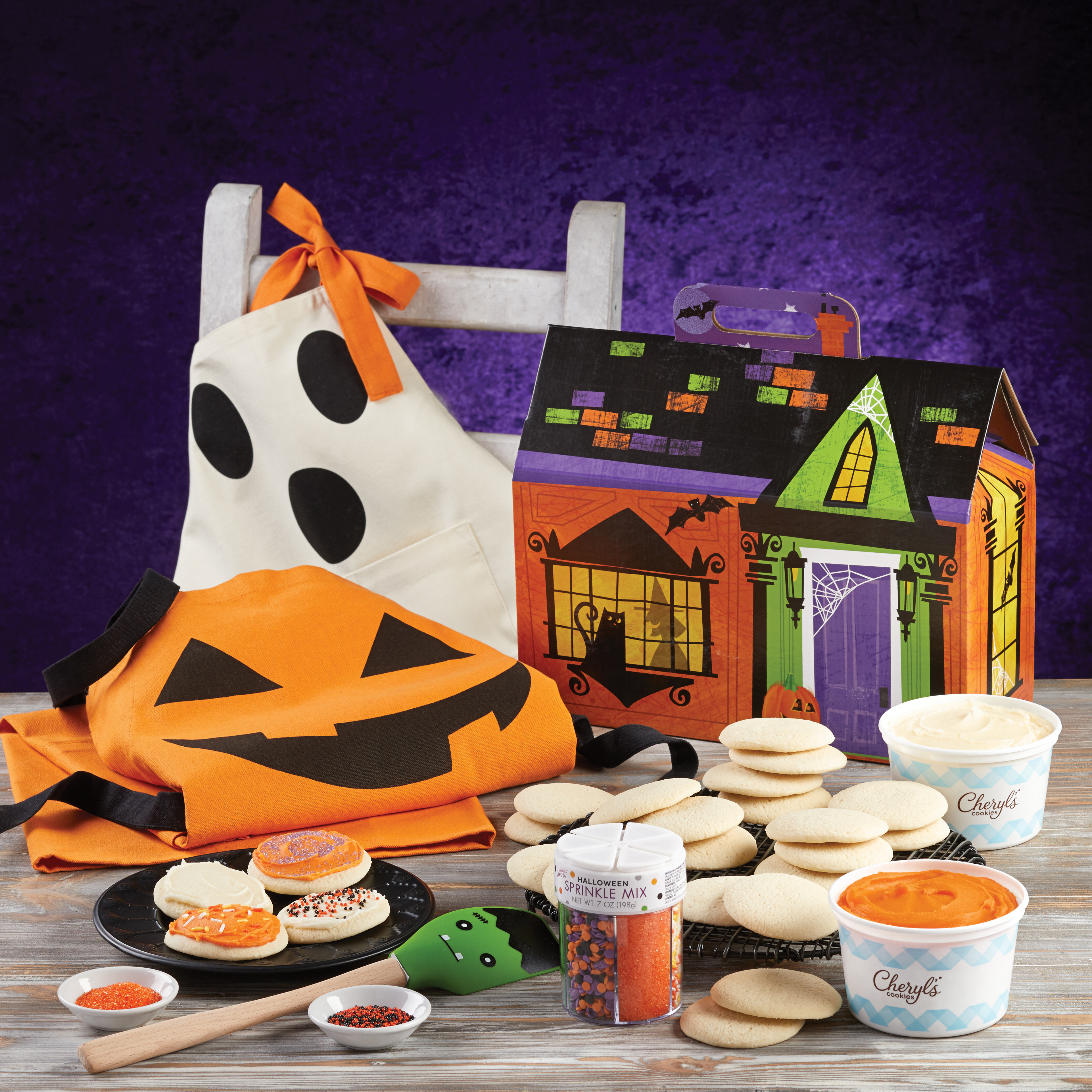 A Halloween display of products from the Cheryl’s Cookies Halloween Cut-Out Cookie Decorating Kit on a white table in front of a dark purple background