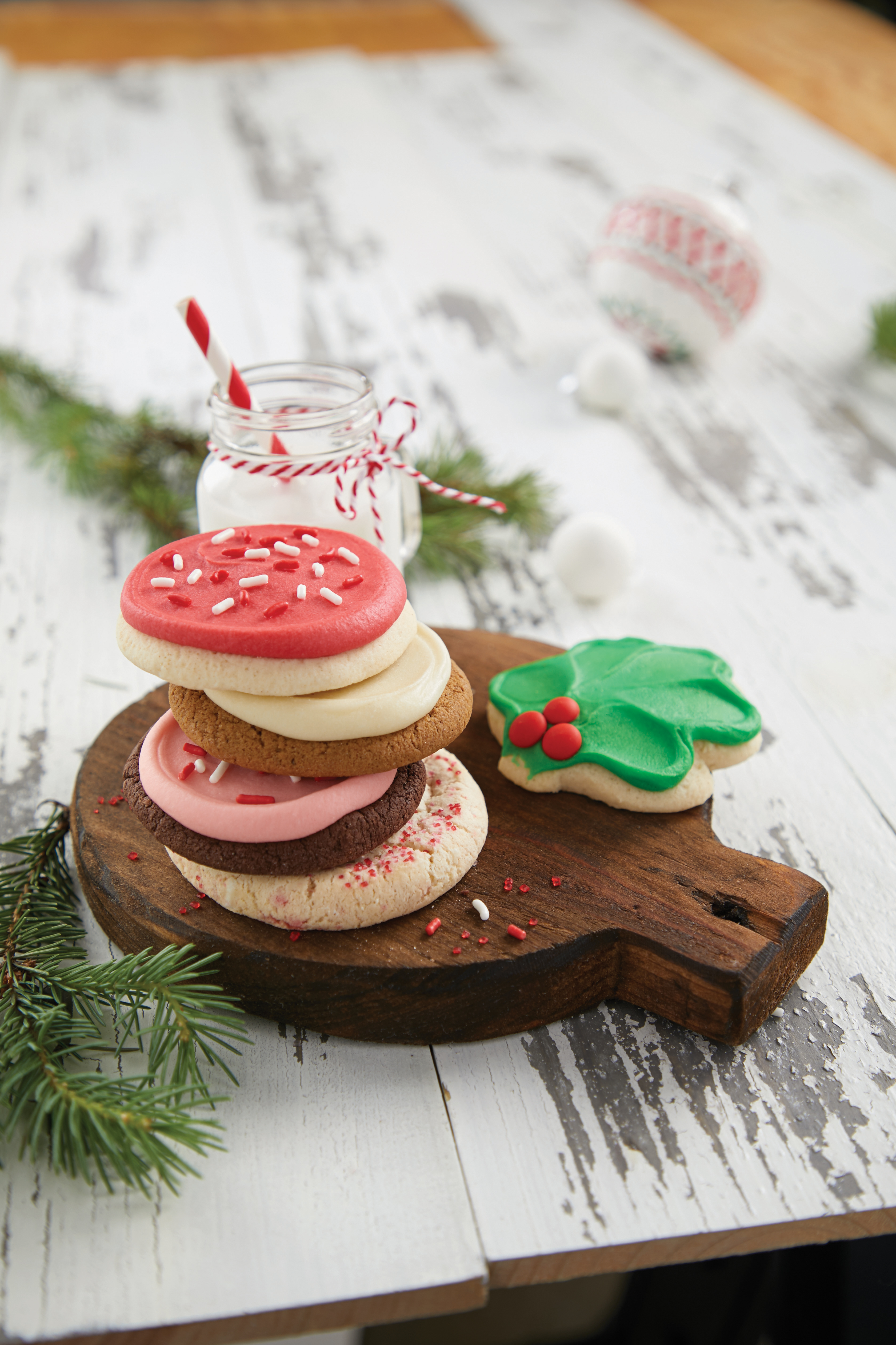 Red, white, green, and pink frosted holiday-themed cookies  from Cheryl's Cookies on a wooden platter next to a glass of milk 