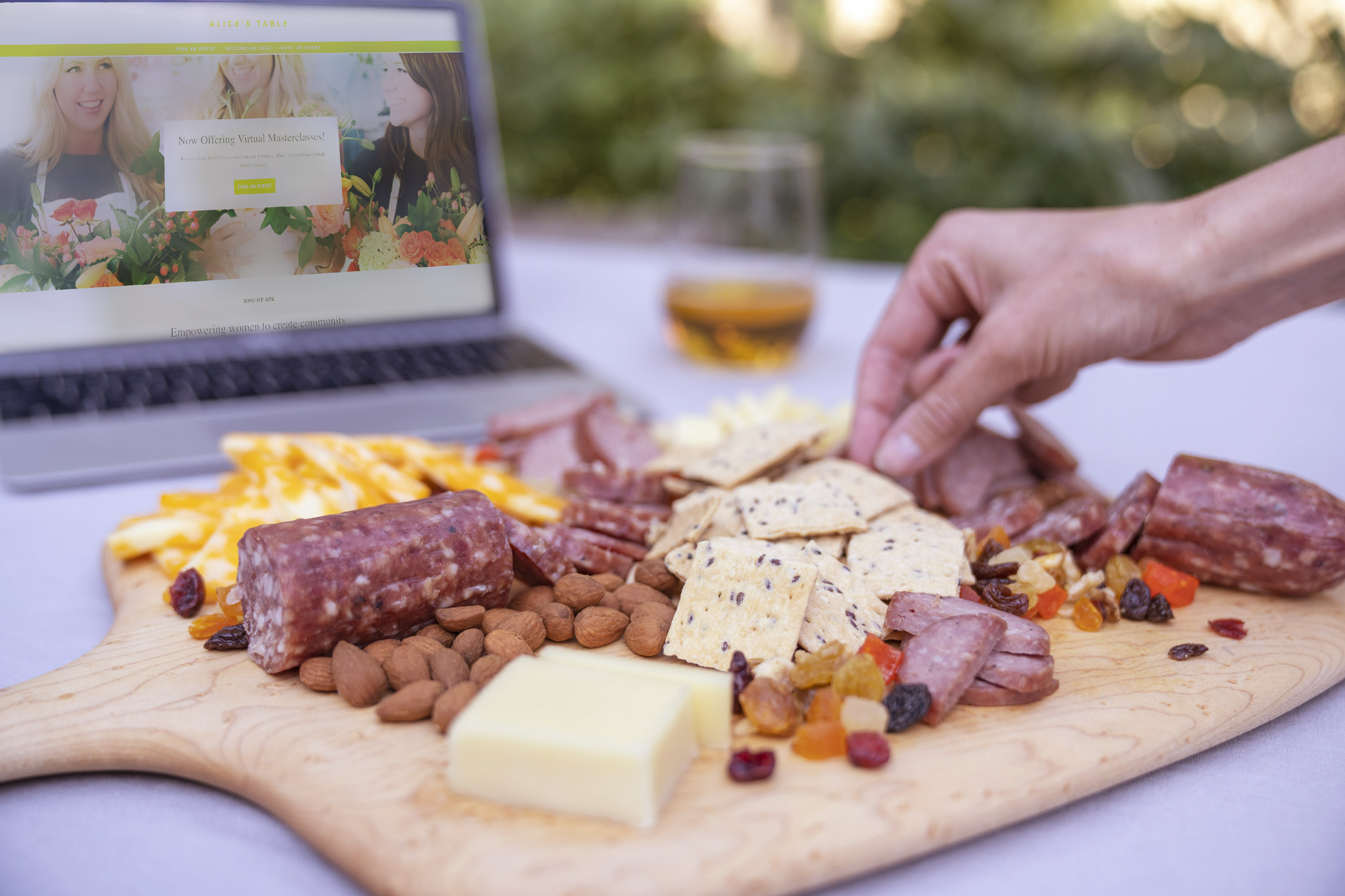 A charcuterie board with gourmet cheese, nuts, cured meats, and dried food next to a tablet that has an Alice’s Table workshop on screen