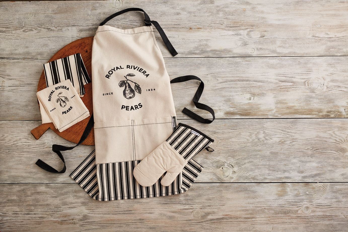 Harry & David Royal Riviera Pears apron and kitchen linens displayed on a brown wooden platter and a white shiplap background
