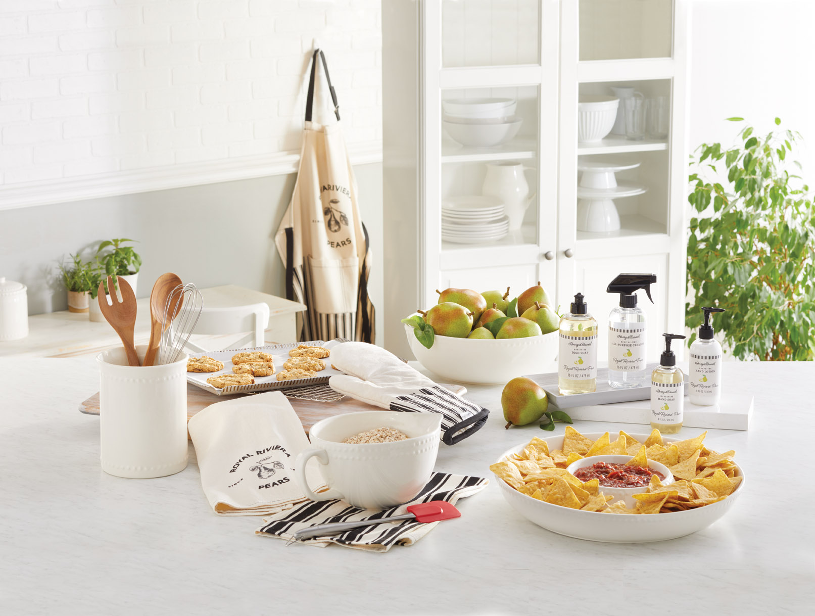 A display of products from the Harry & David Kitchen Collection including linens, hand soap, and more on a clean white kitchen counter with chips in a bowl, cookies on a cooking sheet, and pears in a white bowl