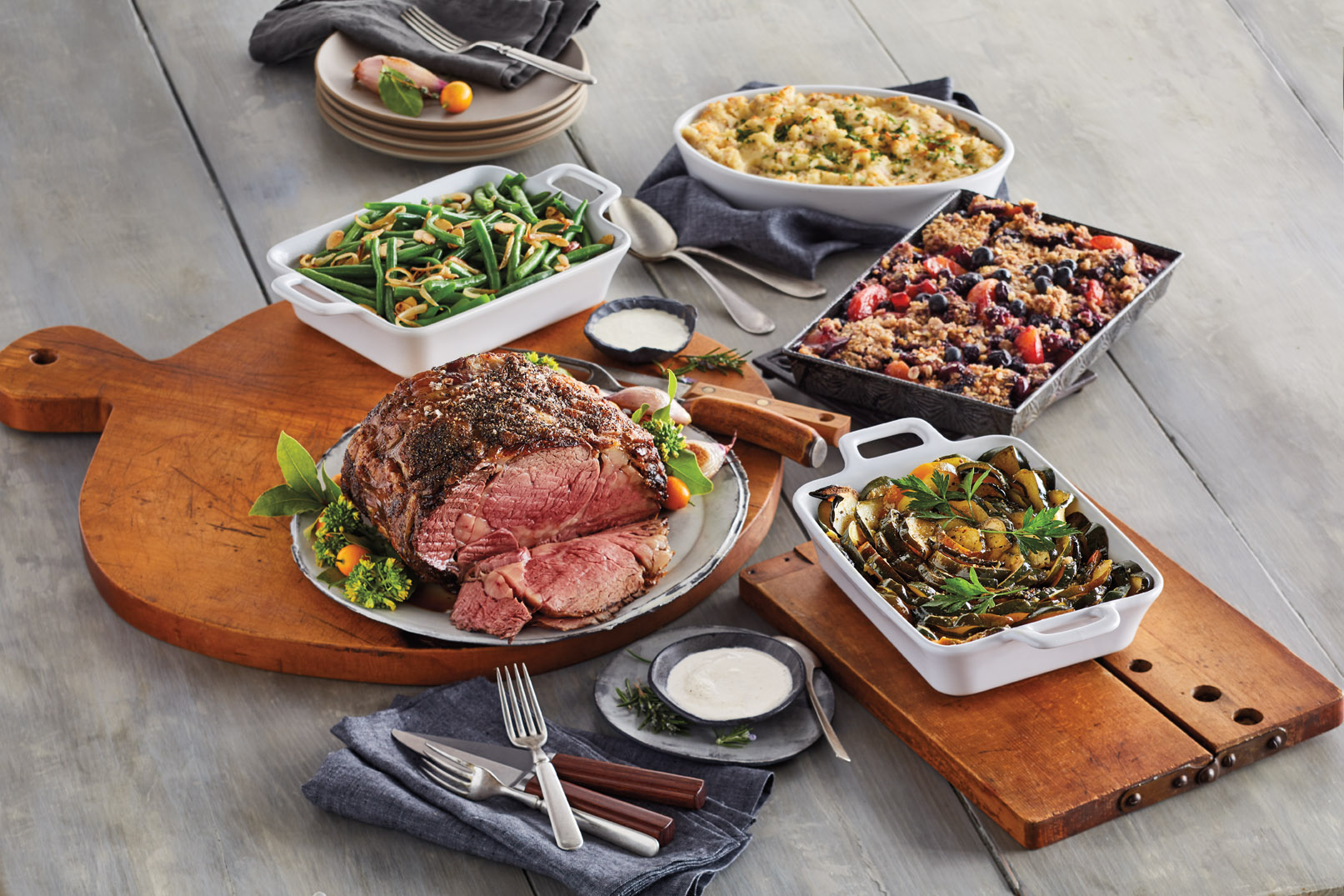 The Harry & David Gourmet Prime Rib Feast with side dishes of mashed potatos and vegetables along with a dessert, accompanyed by navy napkins, light grey plates, and silverware