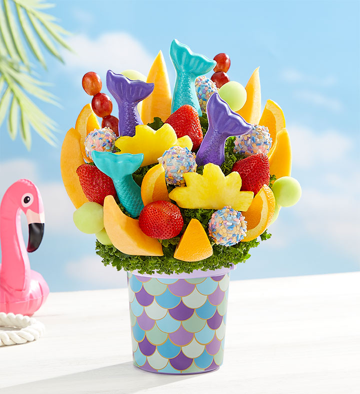A FruitBouquets.com Mermaid Tails Bouquet with mermaid tale-shaped fruit pops and sprinkled cake pops in a purple and blue mermaid-scaled pot