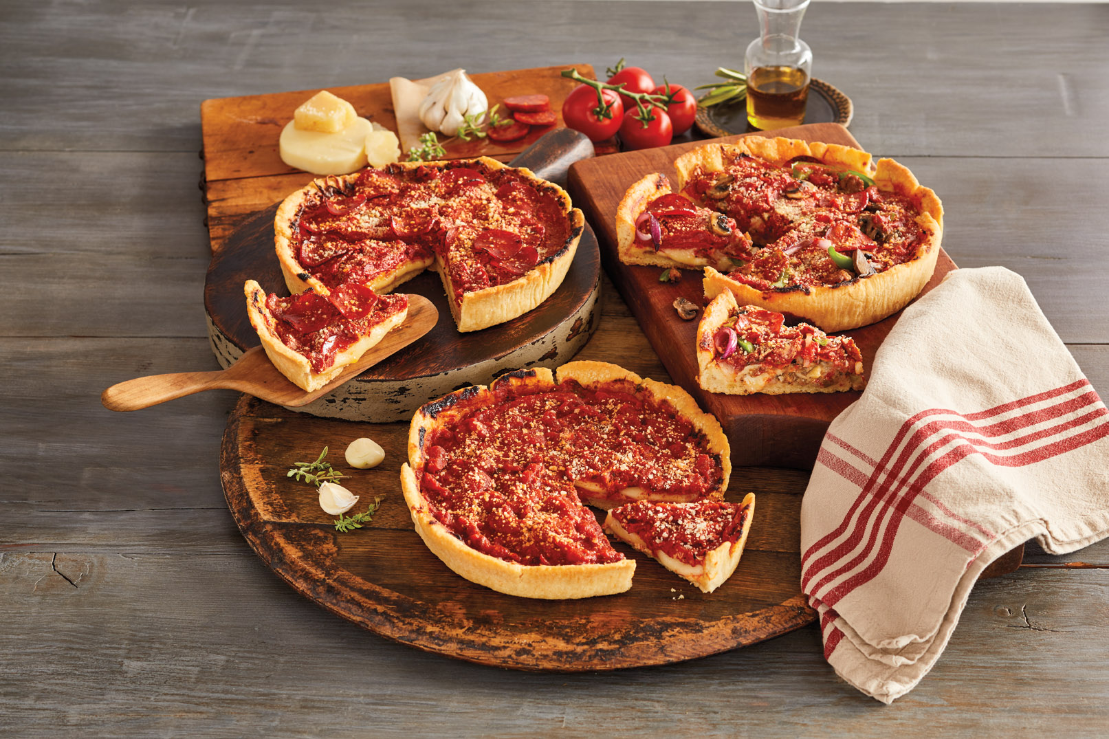 A display of the Pizzeria Uno Original Deep-Dish Pizza Deluxe 3-Pack on wooden platters and garnished with tomatoes, cheese, olive oil, and garlic