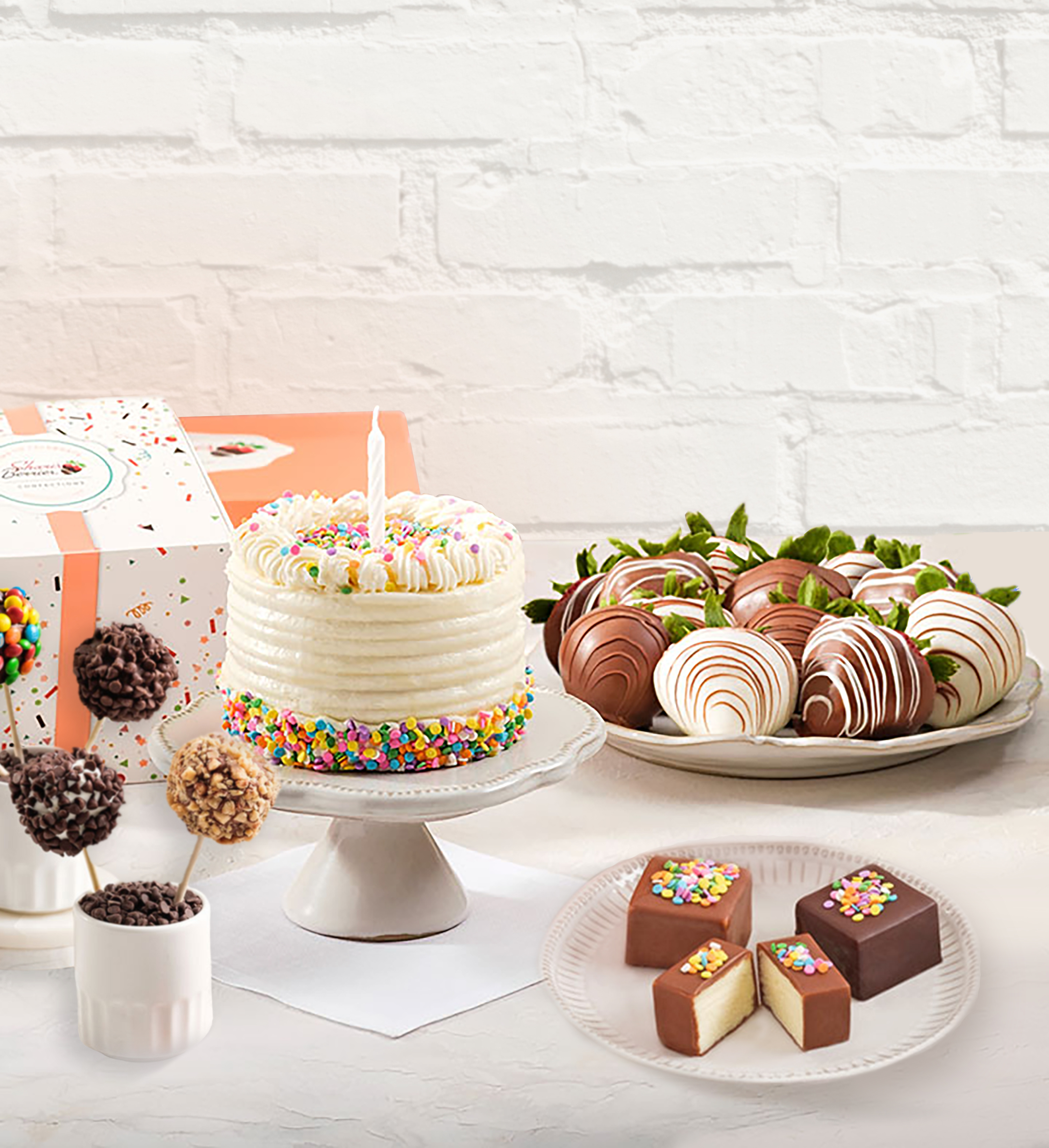An assortment of Shari's Berries Confections, including cake pops, a frosted cake with rainbow sprinkles, chocolate dipped strawberries, and cheesecake bites
