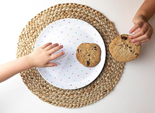 A hand reaching for a chocolate chip cookie on a colorful, polka-dot plate next to another hand holding a chocolate chip cookie